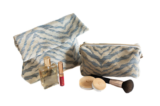 Deux trousses ÉTOFÉ en tissu upcyclé zébré beige et bleu, accompagnées d’accessoires de beauté – pinceau, poudre, parfum, rouge à lèvres.