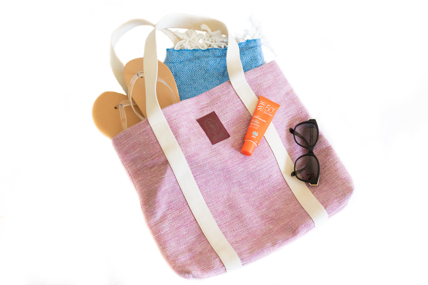 Sac cabas rose ÉTOFÉ en tissu recyclé, rempli d'accessoires d’été : crème solaire, lunettes de soleil, sandales et fouta bleue.
