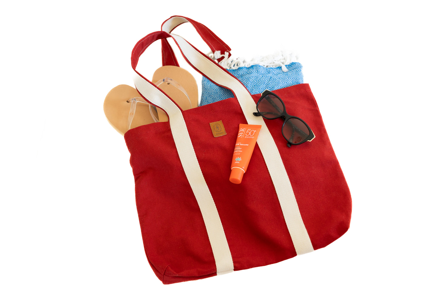 Sac cabas rouge ÉTOFÉ en tissu recyclé, contenant des accessoires d’été : crème solaire, lunettes de soleil, sandales et fouta bleue.