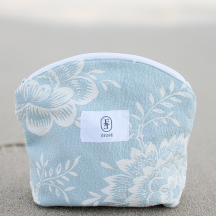 Trousse de maquillage ÉTOFÉ en tissu recyclé bleu clair à motifs floraux, posée sur une plage