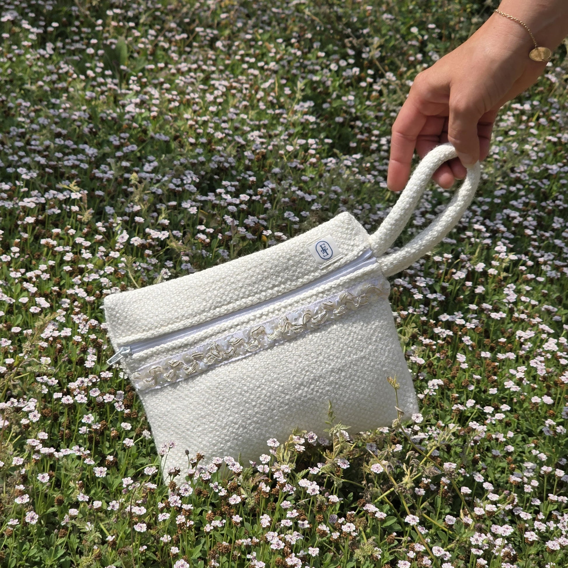 Pochette blanche avec anse tenue à la main au-dessus d’un champ de fleurs champêtre, accessoire éthique pour hôtels et maisons d’hôtes.