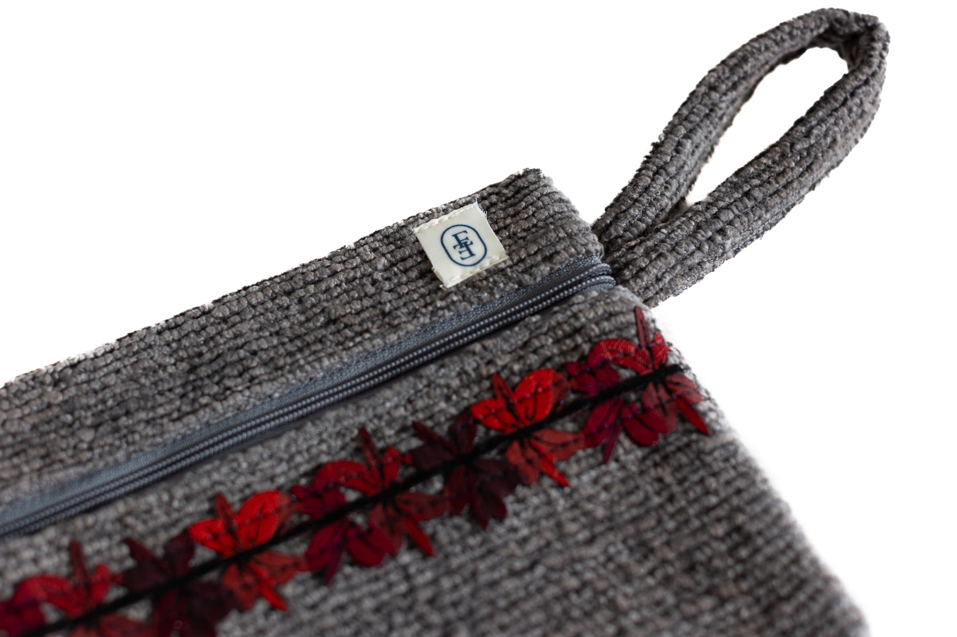Pochette plate ÉTOFÉ grise en tissu texturé upcyclé, ornée d’un galon fleuri rouge et bordeaux, avec fermeture zippée et poignée assortie.