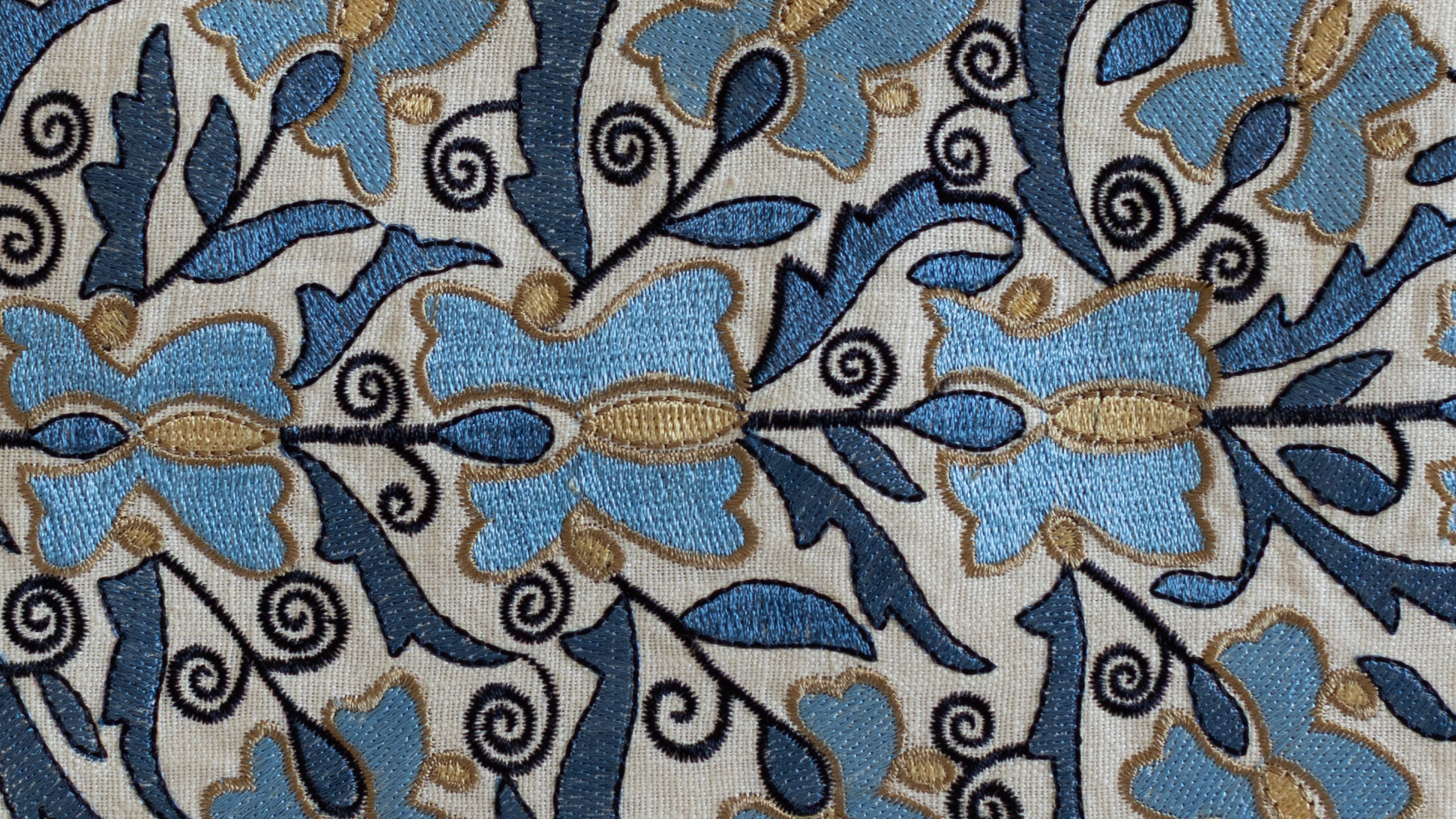 Gros plan sur un tissu brodé représentant des fleurs bleues stylisées avec spirales noires et détails dorés, utilisé dans les créations artisanales d’ÉTOFÉ